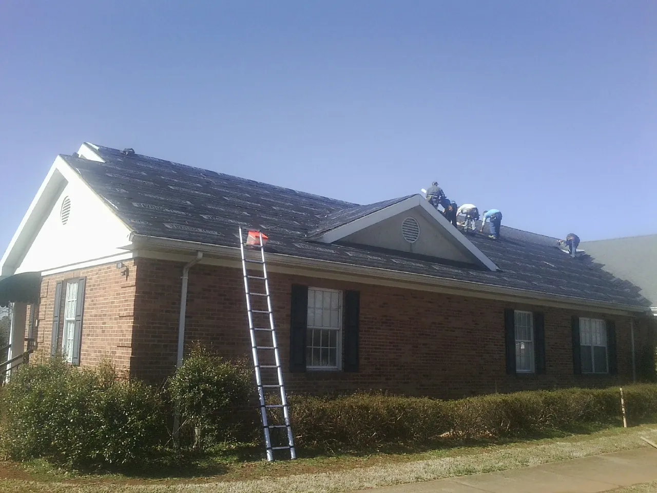Crew tearing shingles off a brick ranch roof with a ladder set — Emergency Roof Repair work by our crew in Loreauville