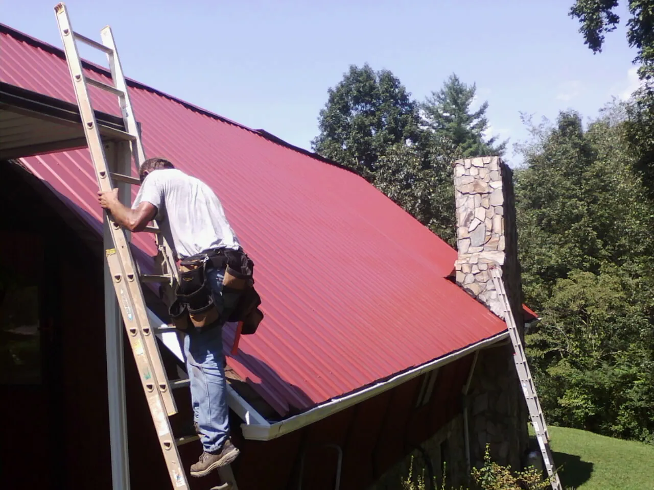 Roofer on a ladder beside a red metal roof and stone chimney — Soffit & Fascia Repair by Roofing Brothers in Loreauville